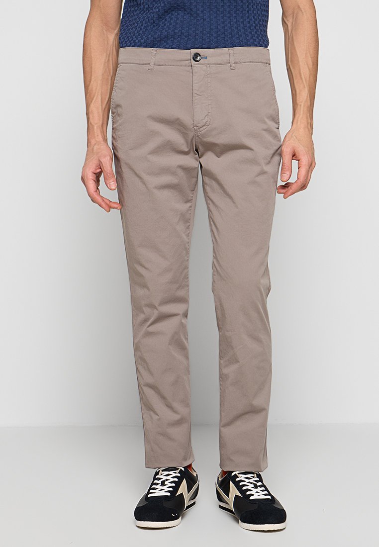 Mann trägt beige, schmal geschnittene Hose, ein blaues Strukturhemd, das in die Hose gesteckt ist, und schwarze Sneakers mit weißen Schnürsenkeln und Akzenten, stehend vor grauem Hintergrund.