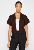 Braune Strickjacke mit kurzen Ärmeln, offenem Vorderteil und strukturiertem Muster. Wird über einem weißen Oberteil getragen und mit schwarzen Hosen kombiniert.