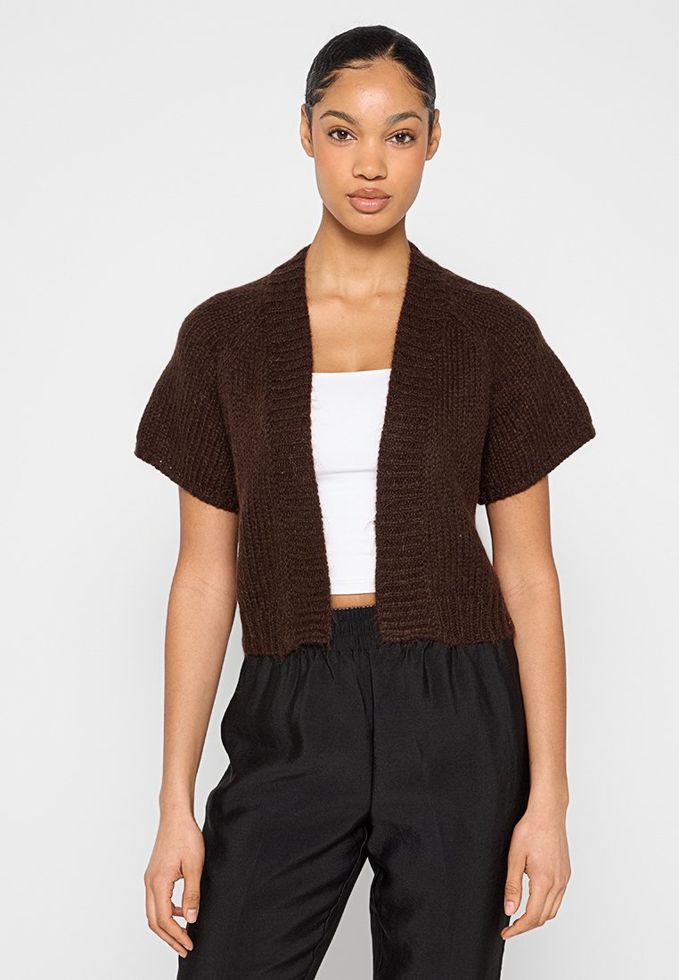 Braune Strickjacke mit kurzen Ärmeln, offenem Vorderteil und strukturiertem Muster. Wird über einem weißen Oberteil getragen und mit schwarzen Hosen kombiniert.