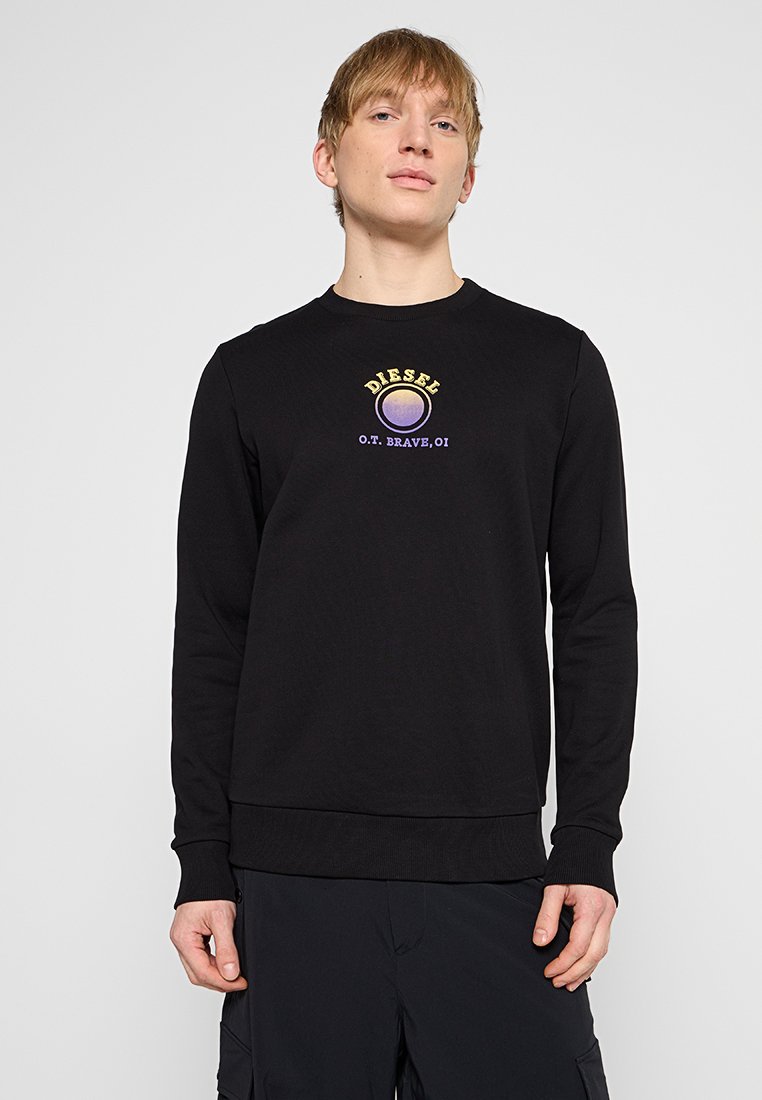 Junger Mann mit hellbraunen Haaren, der ein schwarzes Langarm-Sweatshirt mit einem gelben und lila Diesel-Logo auf der Brust trägt.