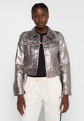 Metallisch silberne Jeansjacke mit einem cropped Design, Fronttaschen und bronzefarbenen Knöpfen, kombiniert mit einem schwarzen Shirt und hellen Hosen.