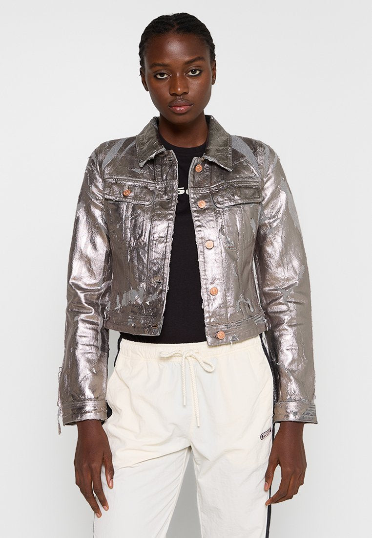 Metallisch silberne Jeansjacke mit einem cropped Design, Fronttaschen und bronzefarbenen Knöpfen, kombiniert mit einem schwarzen Shirt und hellen Hosen.