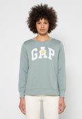 Frau mit lockigem Haar trägt ein hellgrünes GAP-Sweatshirt mit Margeritenmotiv in der Mitte, dazu weiße Hose, steht still.
