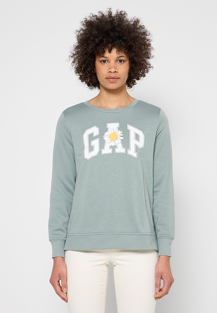 Frau mit lockigem Haar trägt ein hellgrünes GAP-Sweatshirt mit Margeritenmotiv in der Mitte, dazu weiße Hose, steht still.