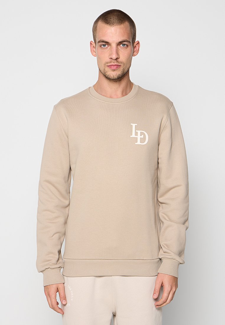 Junger Mann, der einen beigen Sweatshirt mit weißem "LD"-Logo auf der Brust trägt, vor einem schlichten weißen Hintergrund steht.