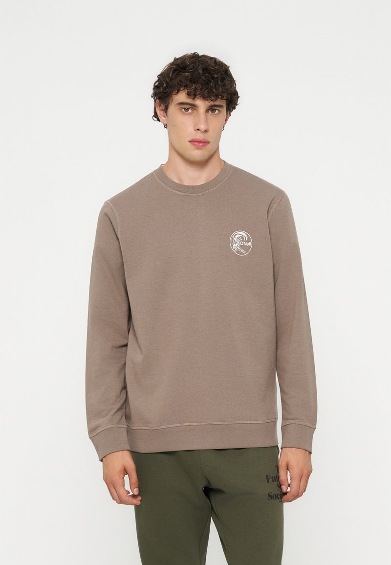 Junger Mann mit lockigem Haar, der einen hellbraunen Langarm-Sweatshirt mit kleinem weißem Logo auf der Brust und dunkelgrünen Hosen trägt.