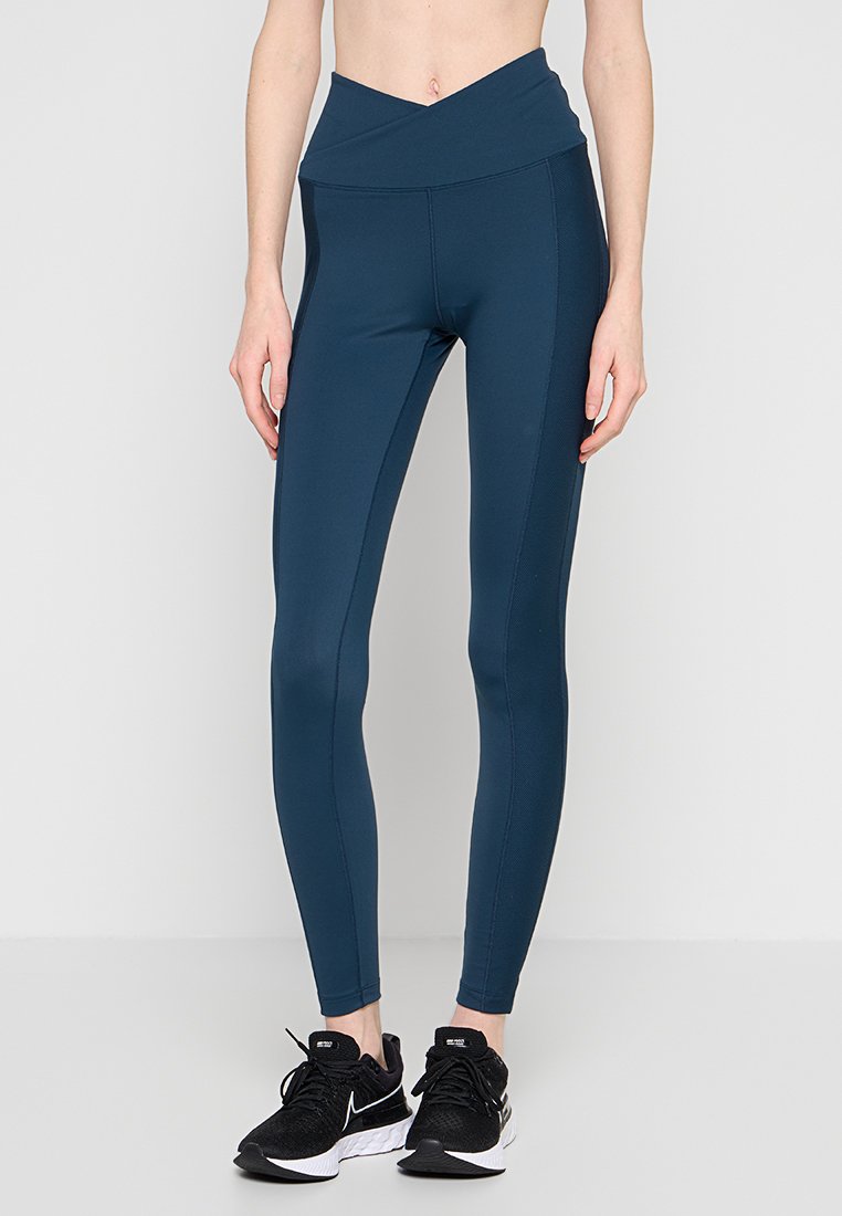 Blaue Leggings mit hoher Taille aus dehnbarem Stoff, mit glatter Textur und Seitennähten für eine enganliegende Optik, kombiniert mit schwarzen Turnschuhen.