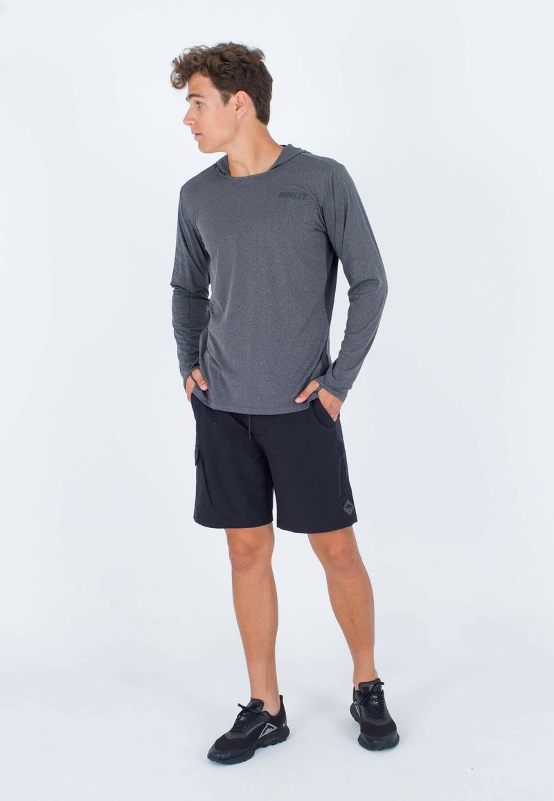 Langärmliges graues Sportshirt mit Kapuze, kombiniert mit schwarzen Cargo-Shorts und schwarzen Turnschuhen. Glatte Textur, funktionales Design.