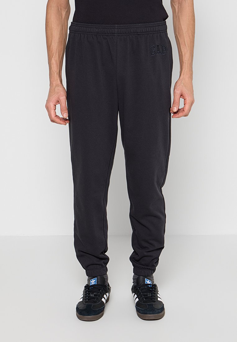 Mann trägt schwarze Jogginghosen mit elastischem Bund und schwarze Adidas-Sneaker, steht vor einem schlichten weißen Hintergrund.