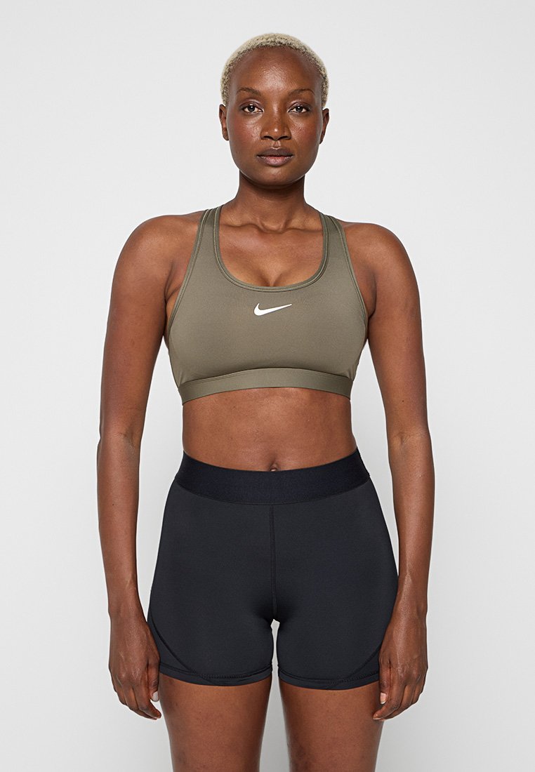 Frau trägt olivgrünen Nike-Sport-BH und schwarze Sportshorts, steht frontal vor hellem einfarbigem Hintergrund.