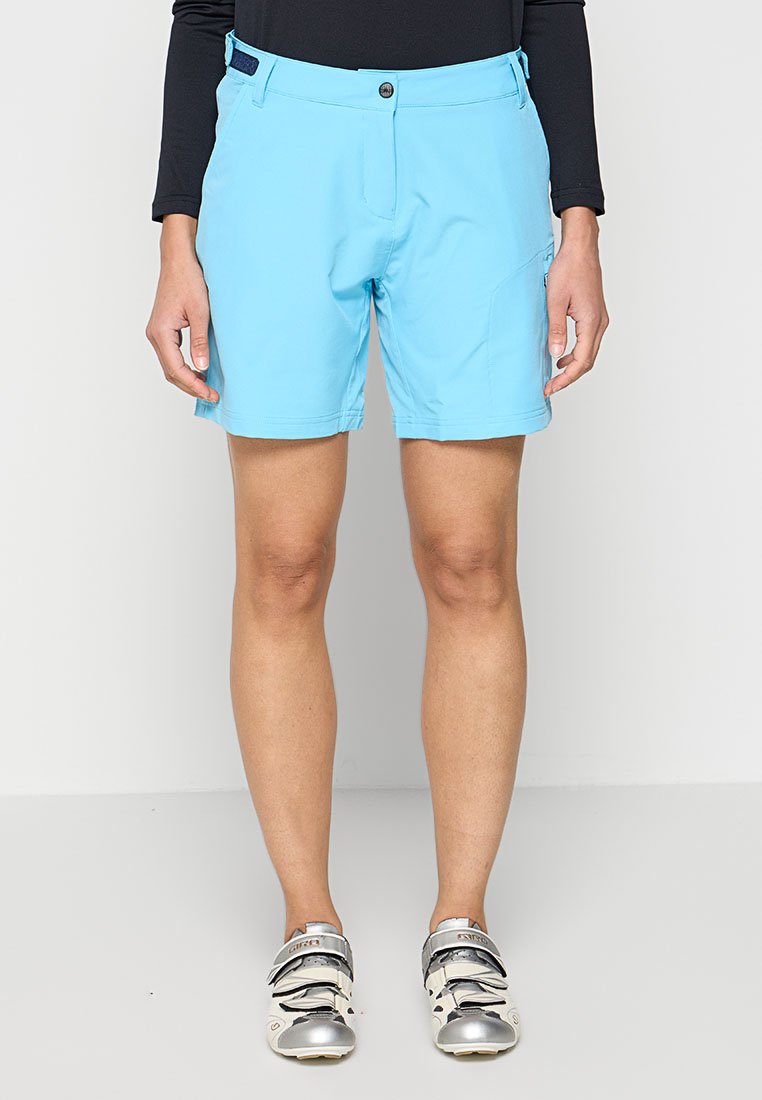 Person in hellblauen Shorts, schwarzem Langarmoberteil und silberweißen Radschuhen steht vor einem weißen Hintergrund.