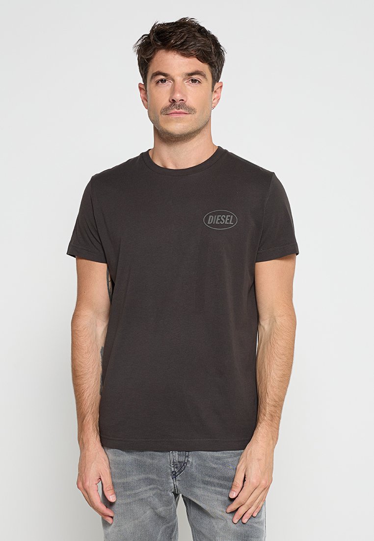 Mann mit Schnurrbart, der ein schwarzes Diesel-T-Shirt und hellgraue Jeans trägt, steht vor einem weißen Hintergrund.