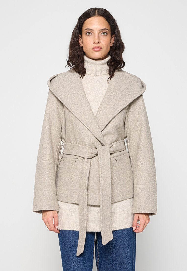Beiger Wollmischungs-Mantel mit breiten Revers, langen Ärmeln und tailliertem Bund. Getragen über einem leichten Pullover und kombiniert mit blauen Jeans.