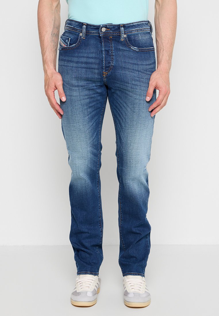 Mann trägt blaue ausgebleichte Slim-Fit-Jeans und weiße Turnschuhe, steht vor einem schlichten hellen Hintergrund.