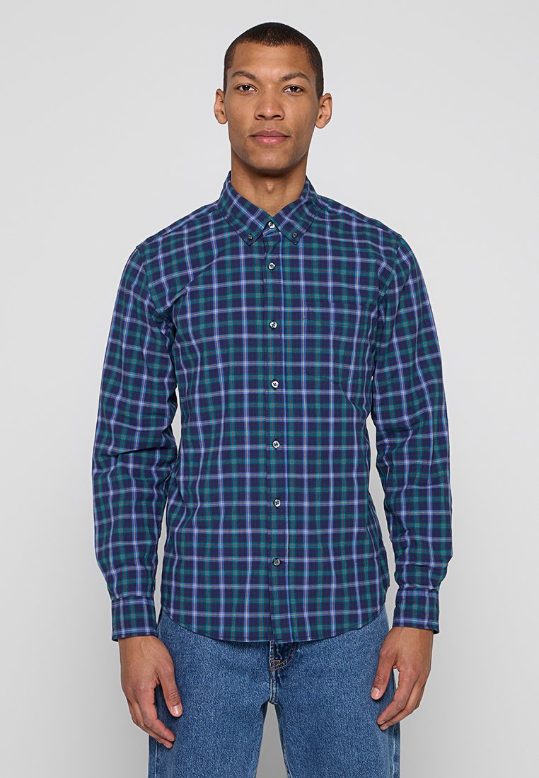 Mann, der nach vorne steht und ein blau-grünes kariertes Hemd mit Knöpfen sowie eine blaue Jeans trägt, vor einem schlichten grauen Hintergrund.