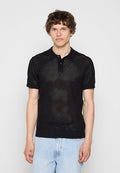 Junger Mann mit lockigem Haar, der ein schwarzes kurzärmeliges Mesh-Poloshirt und hellblaue Jeans trägt, steht vor einem einfarbigen Hintergrund.