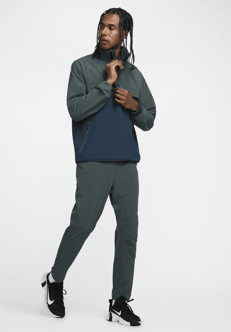 Aktivbekleidungs-Outfit für Herren mit einer zweifarbigen, durchgehenden Jacke in dunkelgrün und navy, kombiniert mit passenden schmal geschnittenen Hosen und schwarzen Sneakers.