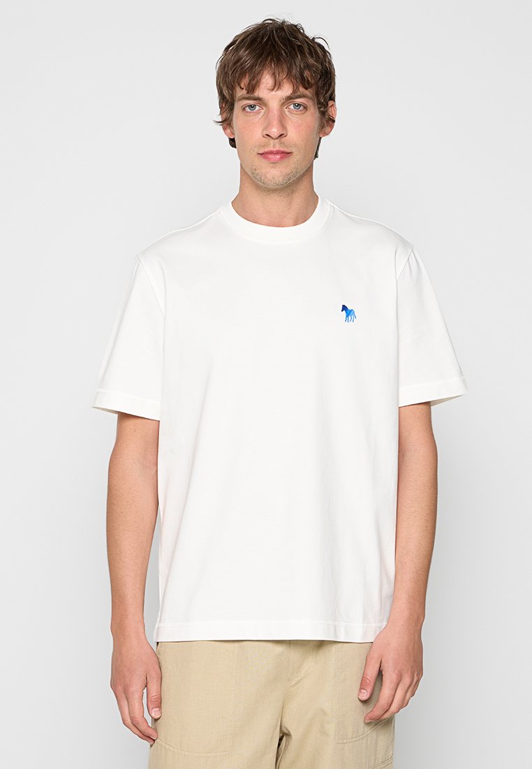 Junger Mann mit kurzen braunen Haaren, der ein schlichtes weißes T-Shirt mit kleinem blauem Pferdelogo und beigen Hosen vor hellem Hintergrund trägt.