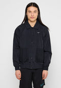 Junge Person mit langen dunklen Haaren trägt eine schwarze Nike-Jacke mit Netz-Taschen und schwarze Nike-Shorts vor weißem Hintergrund.
