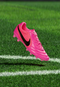 Leuchtend pinke Nike Fußballschuh mit schwarzem Swoosh schwebt über grünem Gras nahe weißen Spielfeldlinien.