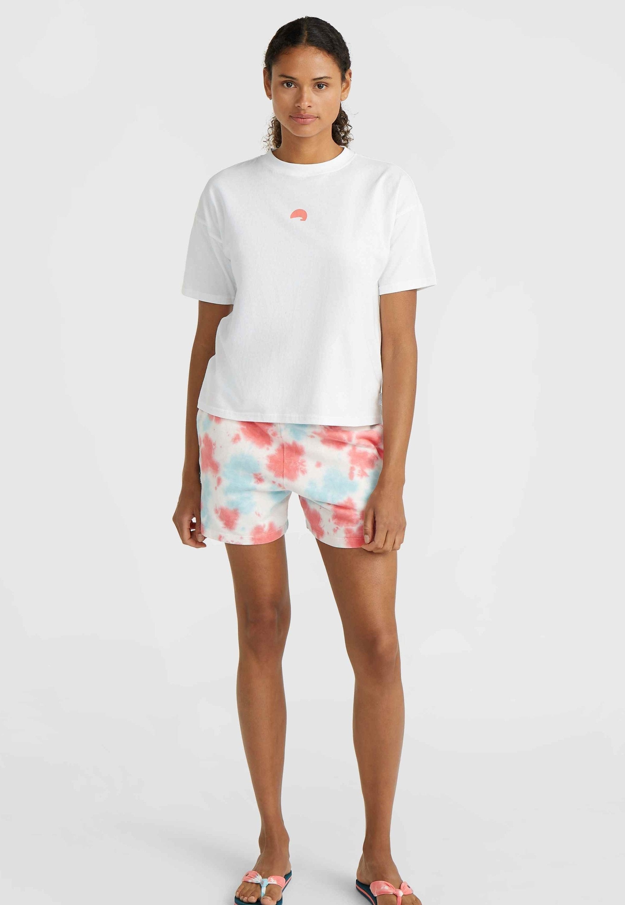 Weißes Baumwoll-T-Shirt mit einem kleinen pinken Halbkreis-Design, kombiniert mit pinken und blauen Batikshorts. Glatte Textur, lässige Passform.