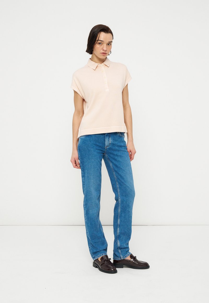 Frau trägt ein hellrosa kurzärmliges Poloshirt, blaue Jeans mit geradem Bein und dunkelbraune Loafers, steht vor einem schlichten weißen Hintergrund.