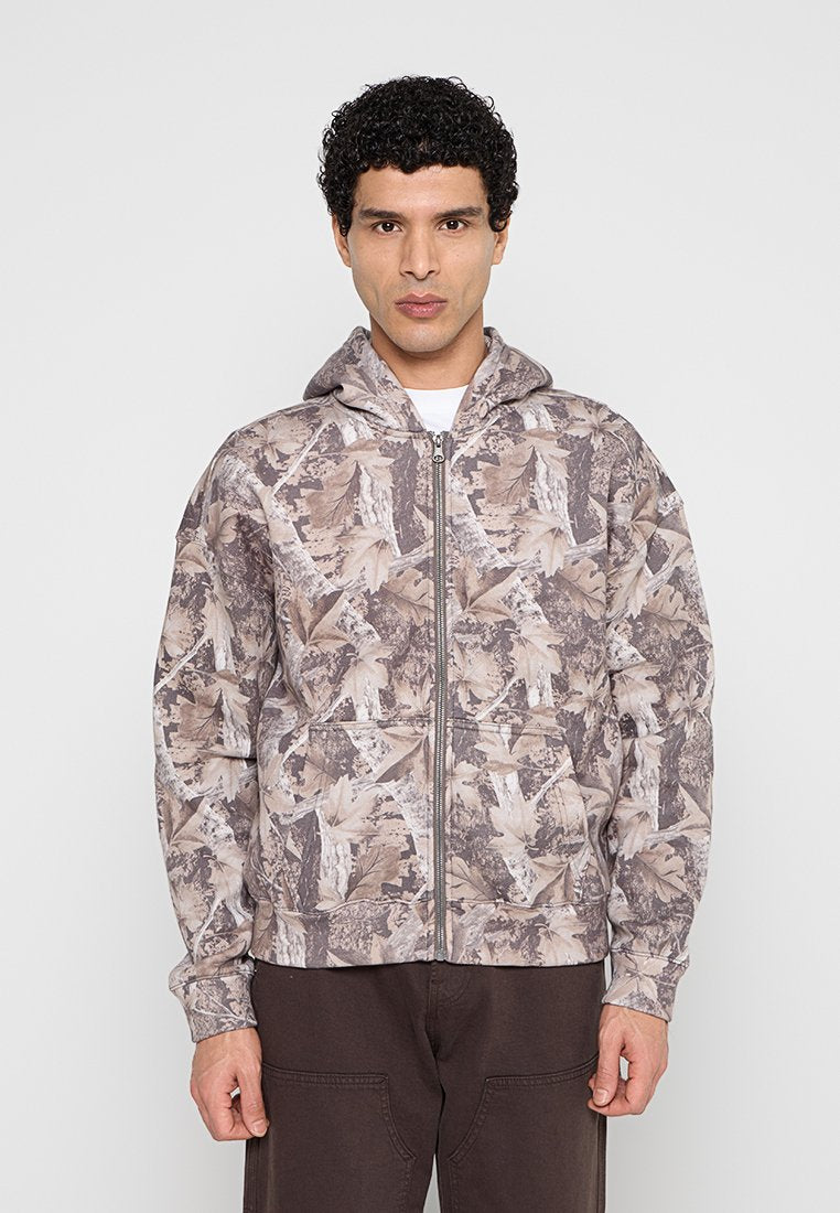 Mann trägt eine braun-beige Jacke mit Blattmuster und Reißverschluss sowie dunkelbraune Hose, steht vor einem schlichten hellen Hintergrund.