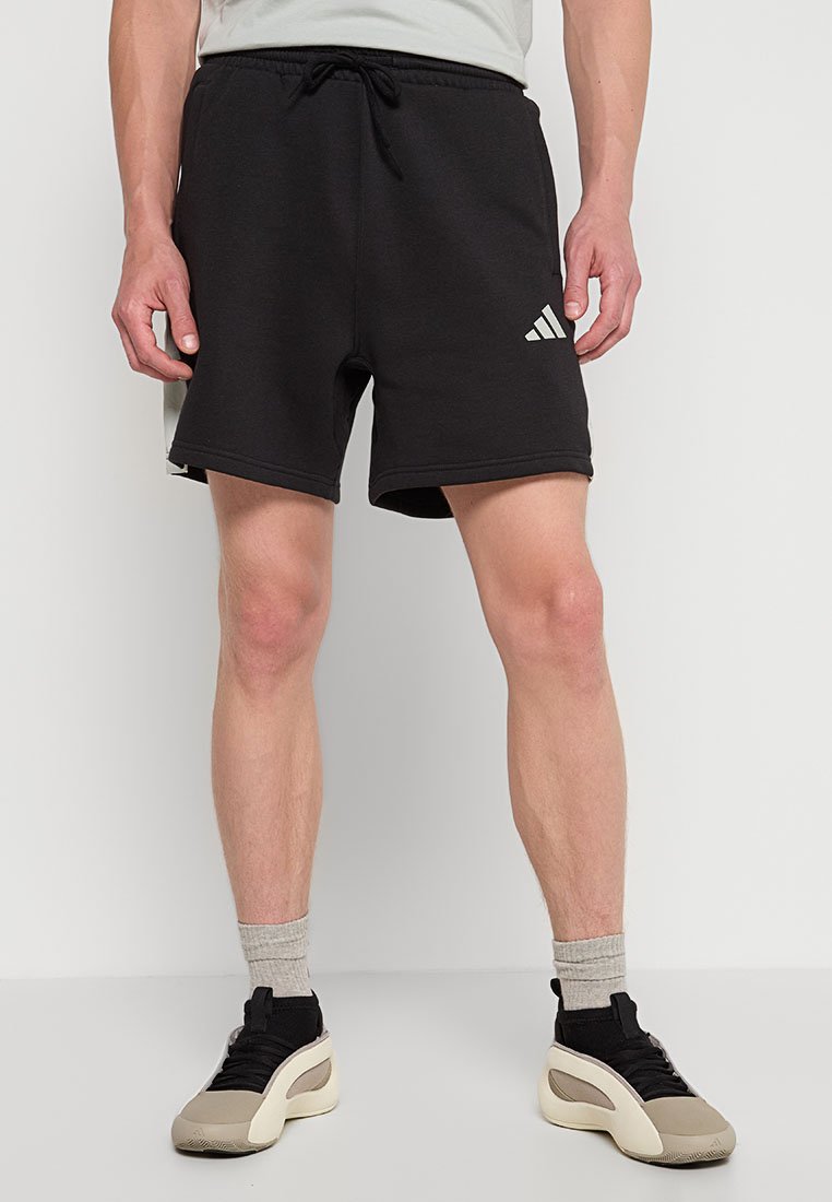 Mann in schwarzen Adidas-Shorts mit Kordelzug, grauen Socken und beigen Sneakers, der vor einem schlichten hellen Hintergrund steht.
