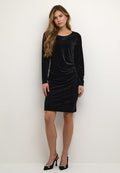Schwarzes samtiges Langarmkleid mit taillierter Silhouette, seitlicher Drapierung und rundem Ausschnitt. Getragen mit High Heels mit goldenen Akzenten.
