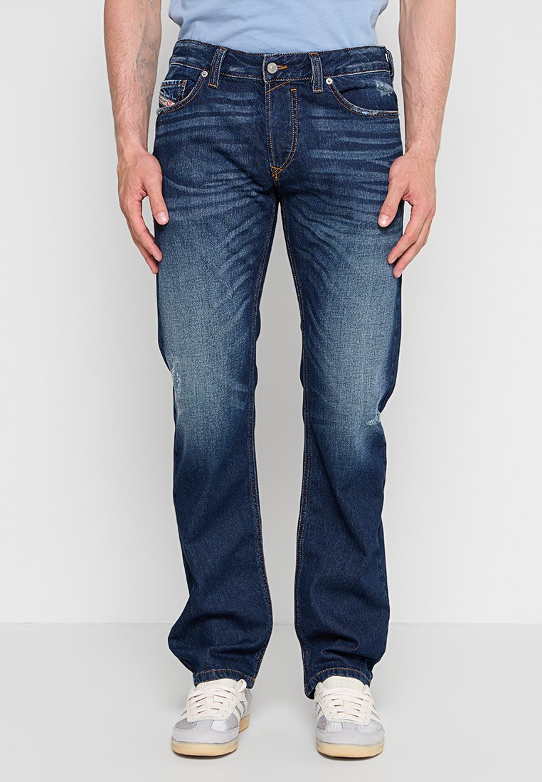 Mann trägt blaue ausgewaschene Jeans mit geradem Bein und weiße Sneakers, steht vor einem schlichten hellen Hintergrund.