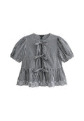 Schwarz-weiße Gingham-Bluse mit kurzen Puffärmeln, drei vorderen Bändern und einer Spitzenborte am Saum. Aus Baumwollstoff, lockere Passform.