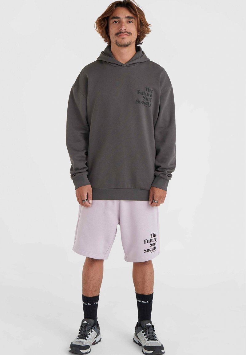 Dunkelgrauer, übergroßer Kapuzenpulli mit einem Logo auf der Vorderseite, kombiniert mit hellrosa Shorts, die dasselbe Logo tragen. Schwarze Socken und graue Sneaker runden das Outfit ab.
