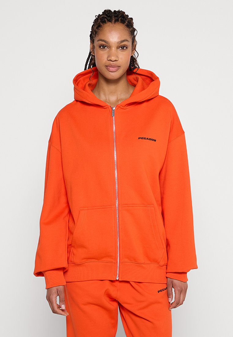 Orangenes Zip-Hoodie mit Kängurutasche, entspanntem Schnitt und subtilen schwarzen Logo-Details auf der linken Seite. Weiches Gewebe.