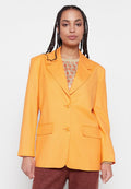 Frau mit geflochtenem Haar, die ein leuchtend orangefarbenes Blazer über einem gemusterten Oberteil und braunen Hosen trägt, vor einem schlichten weißen Hintergrund steht.