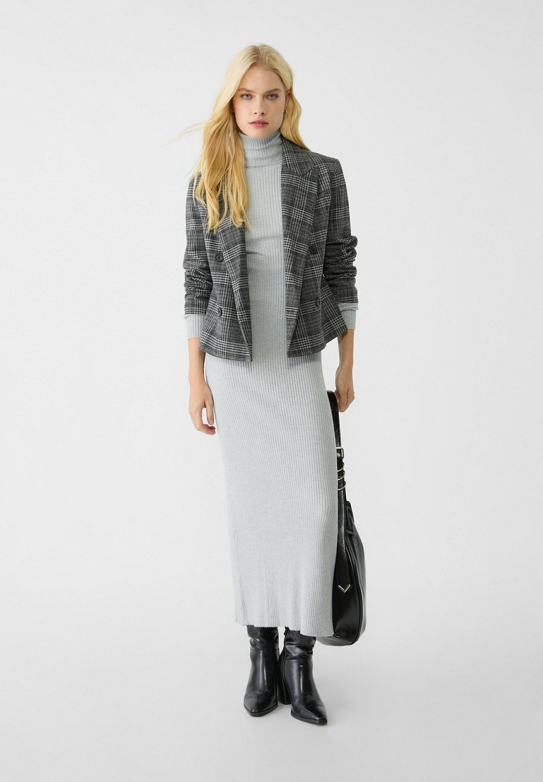 Blonde Frau trägt ein graues, geripptes Rollkragenkleid, einen karierenden Blazer, schwarze Stiefel und hält eine schwarze Handtasche vor einem schlichten weißen Hintergrund.
