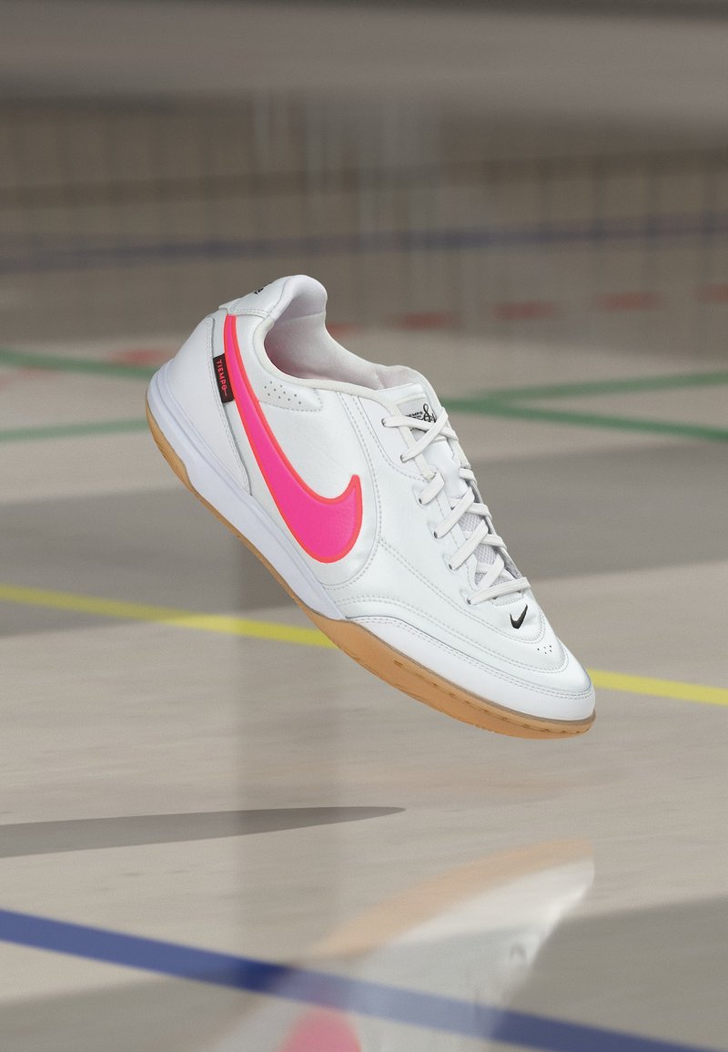 Weißer Indoorschuh für Fußball mit leuchtend pinkem Nike-Swoosh und Gummisohle, in der Luft über einem Hallenboden mit farbigen Linien abgebildet.