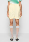 Gelbe maßgeschneiderte Shorts mit hoher Taille und umgeschlagenem Saum, kombiniert mit weißen, knielangen Socken und schwarzen Slippern. Gestreiftes Shirt in Grün und Weiß.