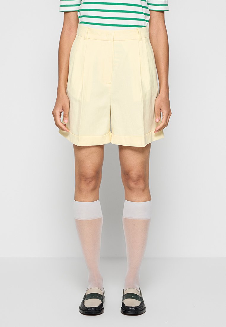 Gelbe maßgeschneiderte Shorts mit hoher Taille und umgeschlagenem Saum, kombiniert mit weißen, knielangen Socken und schwarzen Slippern. Gestreiftes Shirt in Grün und Weiß.