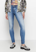 Schlanke, blaue Jeans mit einem verwaschenen Finish, aus Denim gefertigt, kombiniert mit glänzenden silbernen Slippern. Das Outfit besteht aus einem bunten, gemusterten Oberteil und einer bedruckten Jacke.