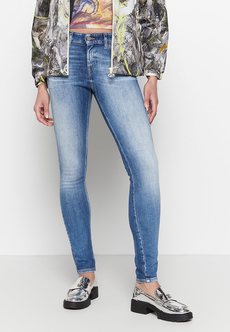 Schlanke, blaue Jeans mit einem verwaschenen Finish, aus Denim gefertigt, kombiniert mit glänzenden silbernen Slippern. Das Outfit besteht aus einem bunten, gemusterten Oberteil und einer bedruckten Jacke.