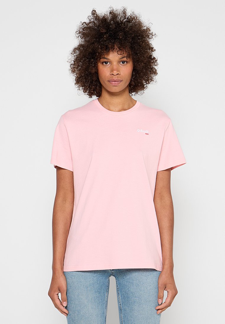 Frau mit lockigem Haar, die ein hellrosa T-Shirt mit kurzen Ärmeln und hellblaue Jeans trägt, steht vor einem schlichten hellen Hintergrund.