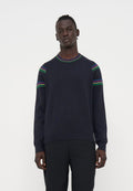 Mann trägt einen dunkelblauen Pullover mit roten, blauen, grünen und gelben Streifen an Schultern und Kragen, kombiniert mit schwarzen Hosen, steht neutral.