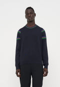 Mann trägt einen dunkelblauen Pullover mit roten, blauen, grünen und gelben Streifen an Schultern und Kragen, kombiniert mit schwarzen Hosen, steht neutral.