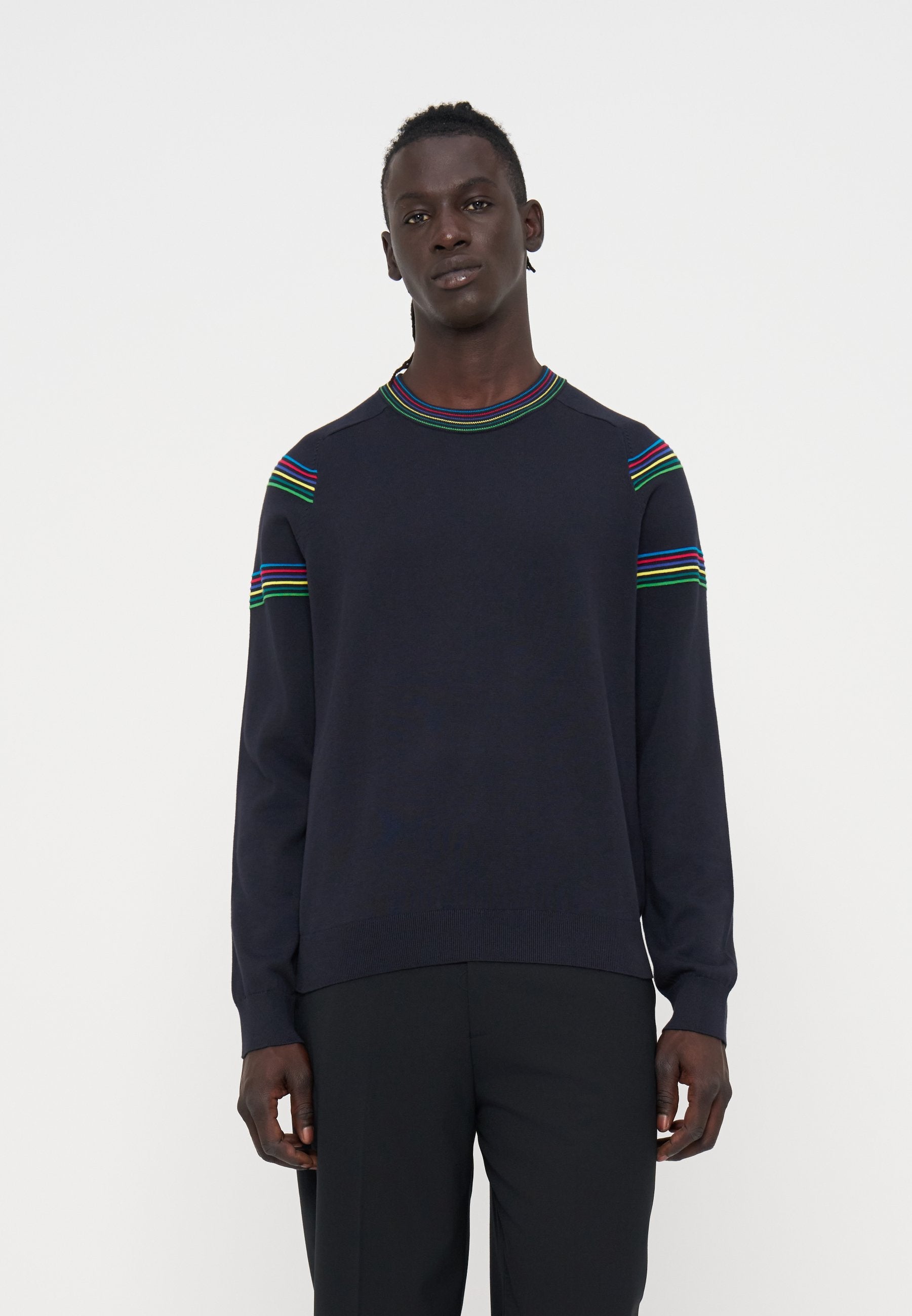 Mann trägt einen dunkelblauen Pullover mit roten, blauen, grünen und gelben Streifen an Schultern und Kragen, kombiniert mit schwarzen Hosen, steht neutral.