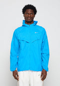 Mann trägt eine hellblaue Nike Windjacke mit Kapuze, steht vor einem einfarbigen hellen Hintergrund und schaut mit neutralem Ausdruck nach vorne.