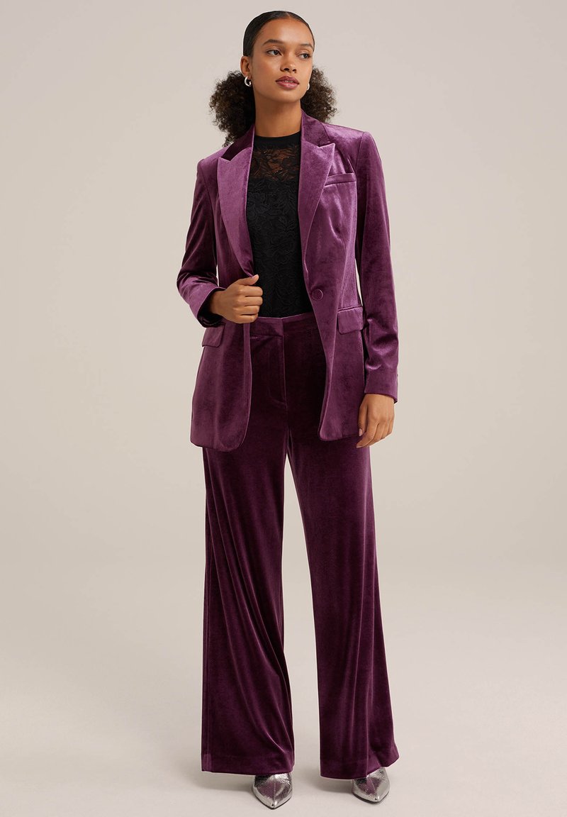 Lila Samtanzug mit einem taillierten Blazer und weit geschnittenen Hosen, kombiniert mit einem schwarzen Spitzentop. Glatte Textur und scharfe Revers.