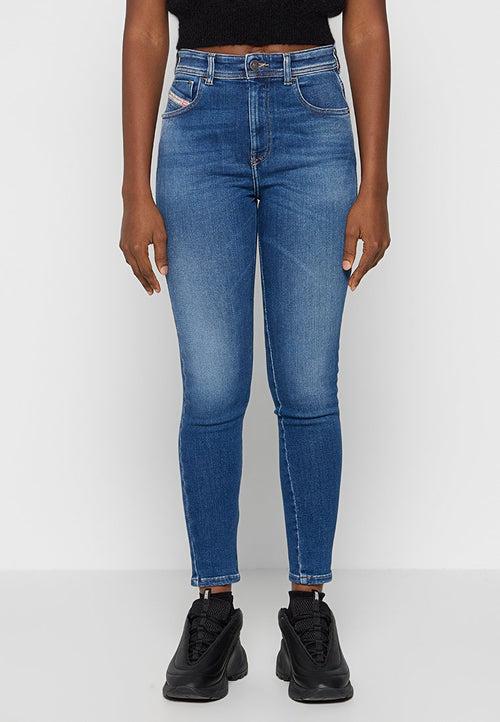 Person, der hochgeschnittene, blaue Skinny Jeans mit Verblendung an den Oberschenkeln und schwarze Sneakers trägt, steht vor einem schlichten weißen Hintergrund.