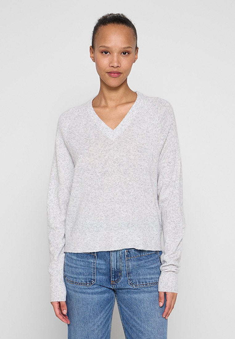 Hellgrauer V-Ausschnittpullover mit langen Ärmeln und gerippten Bündchen, aus einem weichen, strukturierten Stoff. Kombiniert mit blauen Jeans.