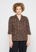 Frau mit lockigem Haar, die eine braune und schwarze Bluse mit Leopardenmuster und Knöpfen trägt, steht vor einem schlichten hellen Hintergrund.