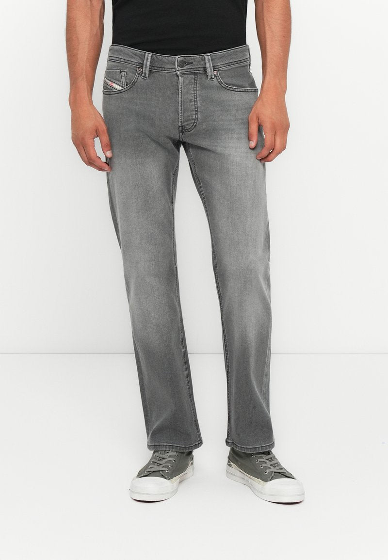 Mann, der verblasste graue geradlinige Jeans, ein schwarzes Shirt und graue Segeltuchschuhe trägt und vor einem weißen Hintergrund steht.